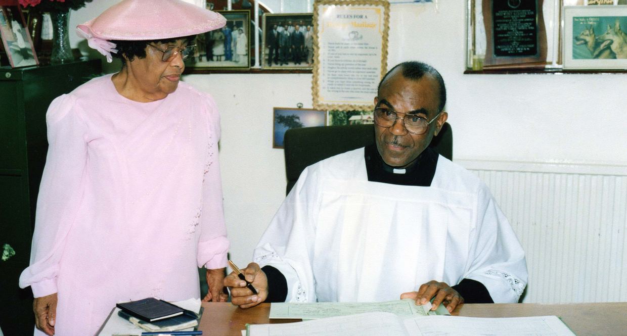 Bishop Dunn in a white robe signing a register at his desk, with Mother Dunn standing beside him