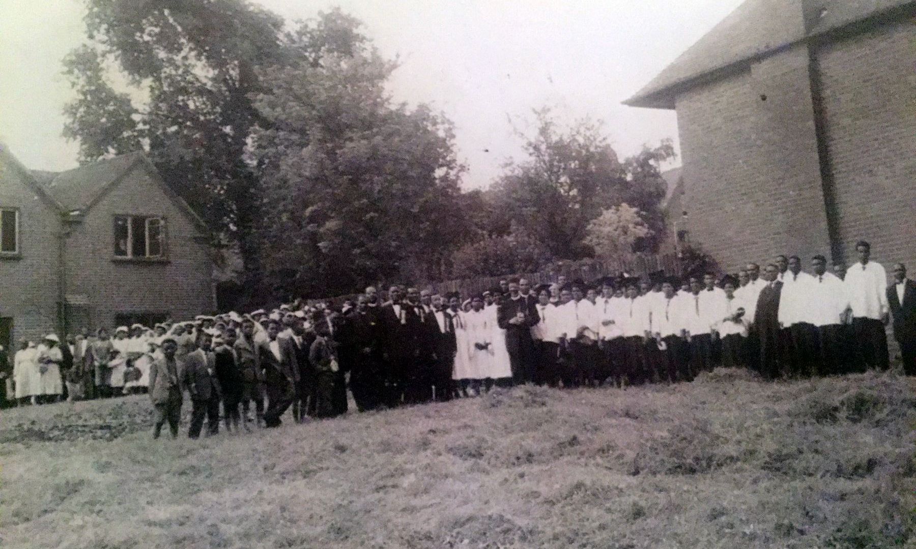 A large congregation, many in white, standing in a line on open ground beside brick houses